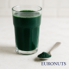 Ποτήρι smoothie με Σπιρουλίνα (Spirulina Powder) σε λευκή μαρμάρινη επιφάνεια, δίπλα κουταλάκι με σκόνη Σπιρουλίνας – EURONUTS Πάτρα